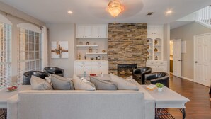 A modern living room with a stone fireplace, white cabinetry, and a dining area with a white table and black leather chairs. - Family Friendly Spacious Home with Backyard Pool by Redawning (Frisco)