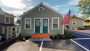 A small, single-story building with a green door and windows, a small porch, and an American flag. - The Lakeview Cottage 9 (Laconia)