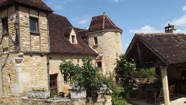 Un edificio de piedra con una torre, una estructura de madera con techo de tejas y un jardín con plantas y flores.