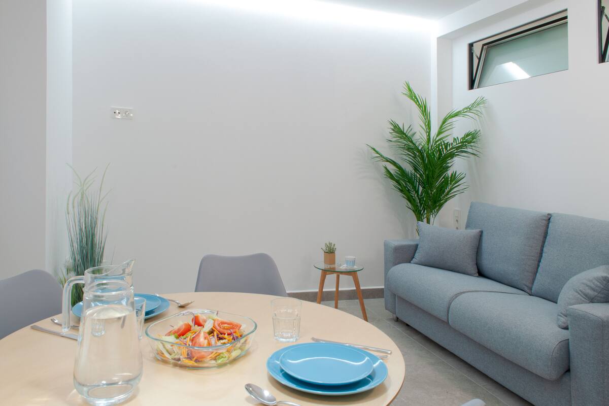 A dining area with a round table, blue plates, a pitcher, and a bowl of salad. There is a grey sofa and a small plant in the corner.