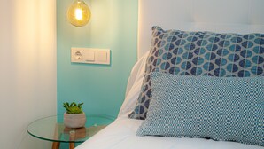 Ein ordentlich bezogenes Bett mit gemusterten Kissen, einer Nachttischlampe und einem kleinen Topf mit Pflanze auf einem Glastisch.
