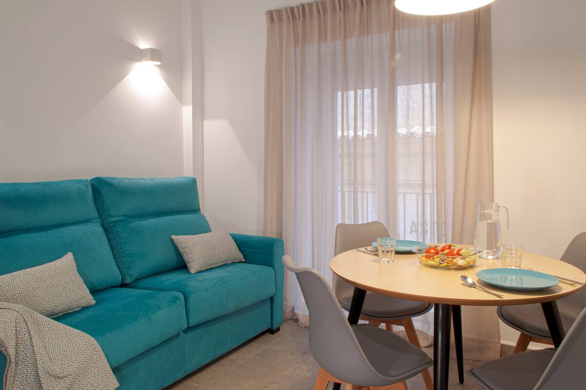 A teal sofa with pillows, a round wooden dining table with chairs, a pitcher, and a bowl of salad.