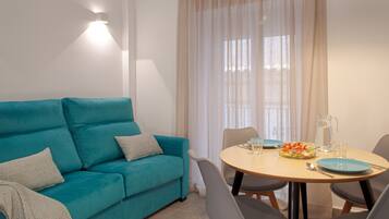 Un sofá azul verdoso con almohadas, una mesa redonda de madera con sillas, una jarra y un tazón de ensalada.