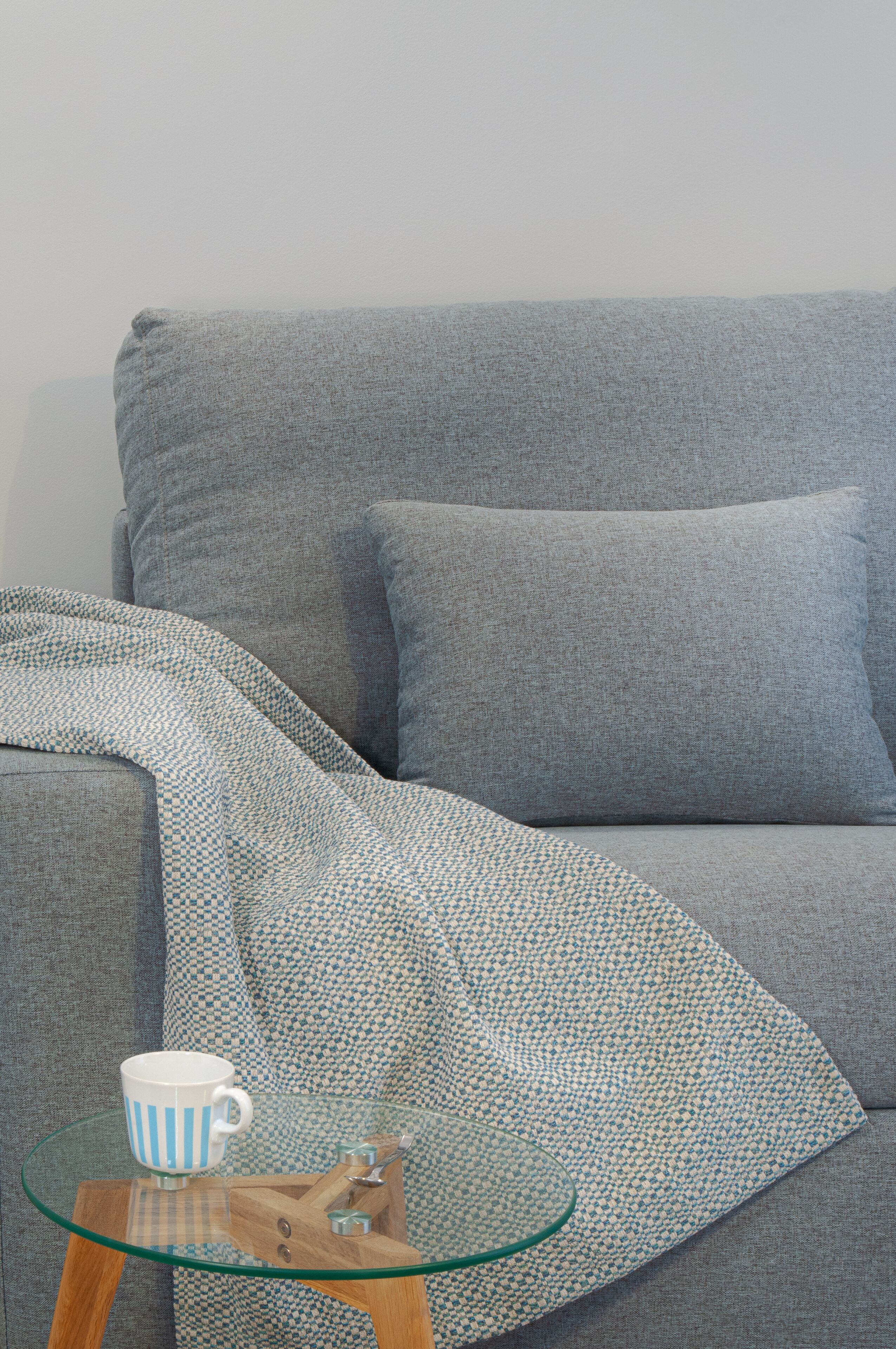 A grey sofa with a patterned throw blanket and a glass coffee table with a blue and white mug.