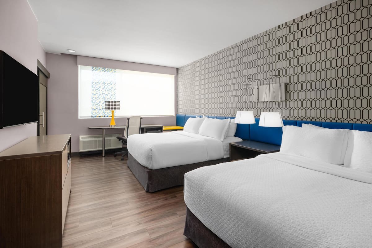 A hotel room with two beds, a desk, a chair, a TV, and a patterned wall.