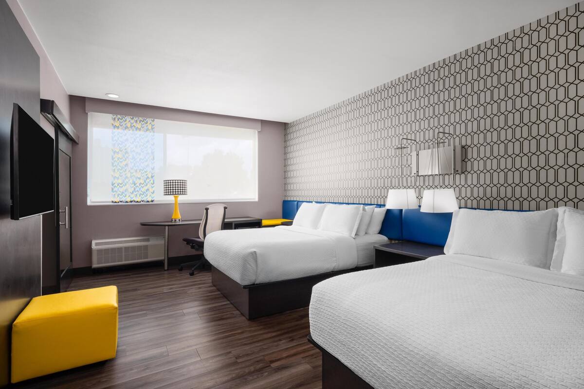 A hotel room with two beds, a desk, a chair, a TV, and a window with patterned curtains.