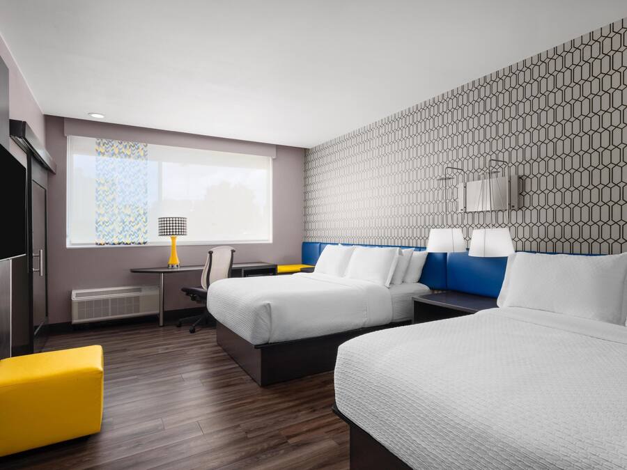 A hotel room with two beds, a desk, a chair, a TV, and a window with patterned curtains.