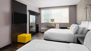 Une chambre d’hôtel moderne avec deux lits, une télévision, une petite table et un pouf jaune.