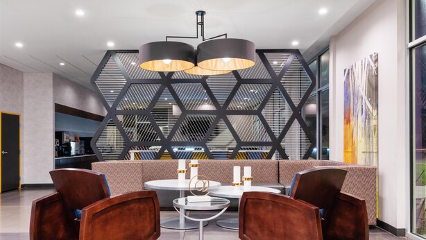 Een moderne lobby met een geometrisch wandontwerp, een zithoek met ronde tafels, en een groot raam dat uitzicht biedt op de omgeving.