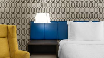 Habitació d'hotel amb paper pintat de dibuixos geomètrics, un panell frontal blau, un llit blanc, un otomà groc i una taula de nit amb una làmpada.