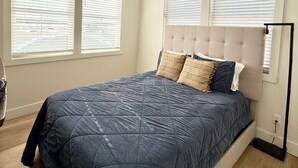 Ein Schlafzimmer mit einem Bett, zwei Fenstern mit Jalousien, einer Deckenleuchte und einer Stehlampe.