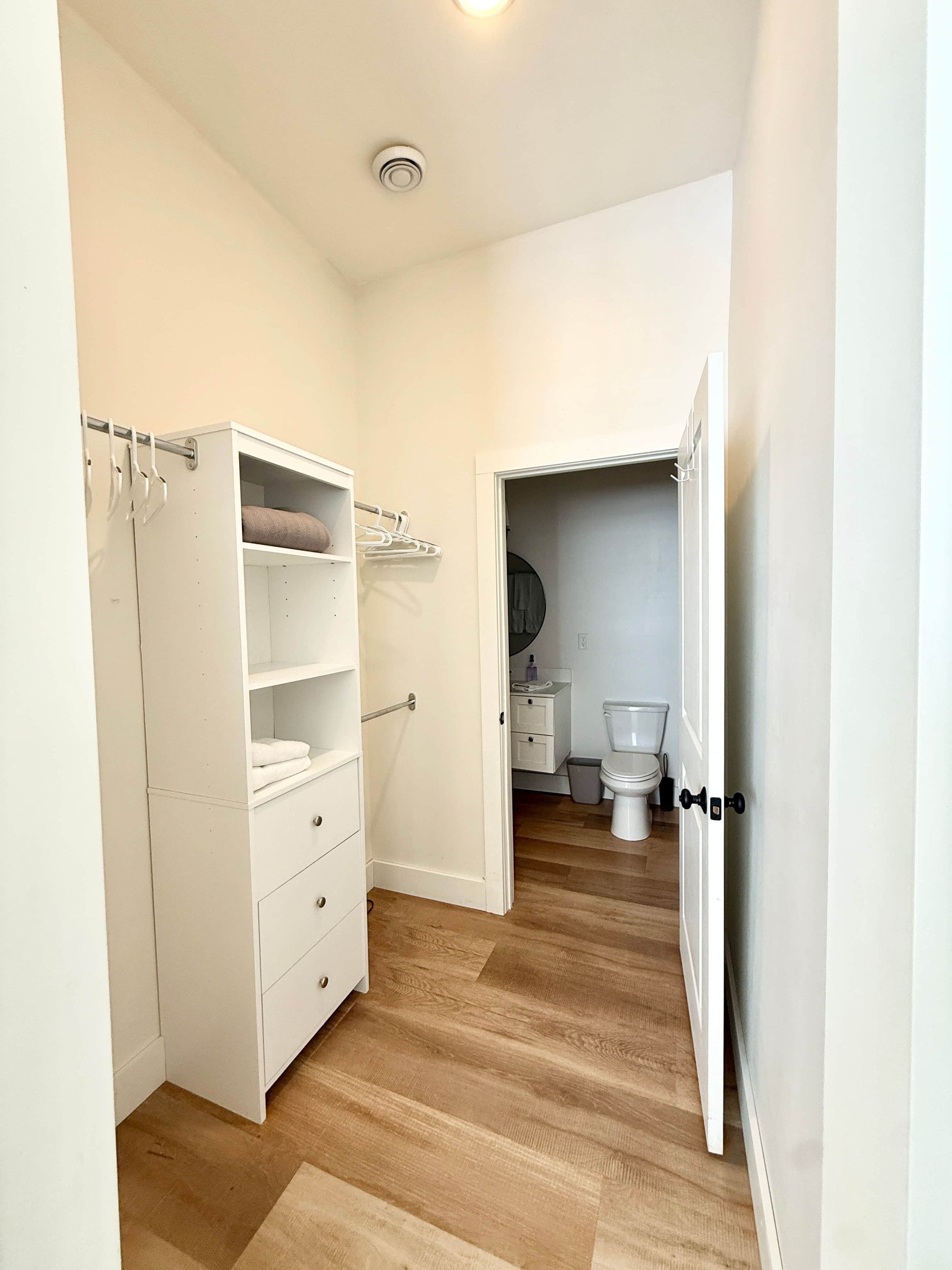 Un couloir étroit avec une armoire blanche, une petite salle de bain avec des toilettes et un lavabo, et un plancher en bois.