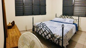 Um quarto com estrutura de cama em metal, mesa de cabeceira de madeira, duas ventoinhas fixadas na parede e duas janelas com persianas.