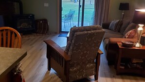 A living room with a wooden floor, a patterned armchair, a sofa, a television, and a sliding glass door leading to an outdoor area. - Nice Condo At Attitash Mountain Village, Close to Storyland, Location! (Bartlett)