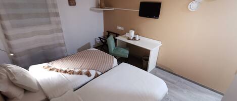 A hotel room with two beds, a desk with a chair, a TV mounted on the wall, and a small shelf with a lamp.