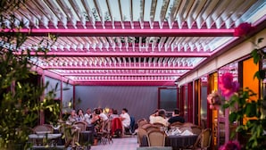 An indoor dining area with tables and chairs arranged under a glass ceiling with pink lighting.