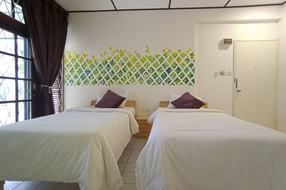 A hotel room with two beds, a window with curtains, a wall with a green and white pattern, and a door with a light switch.