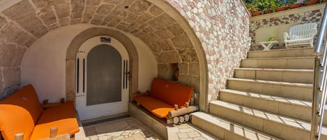 Un espace extérieur aménagé avec des murs en pierre, comprenant un canapé et des coussins, un escalier et une porte dotée d’un panneau de sécurité.
