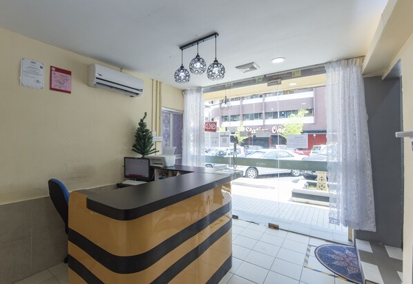 A reception desk with a computer, a small plant, and a window with a view of parked cars and a building outside. - OYO 90254 D Anugerah Hotel (Malacca City)
