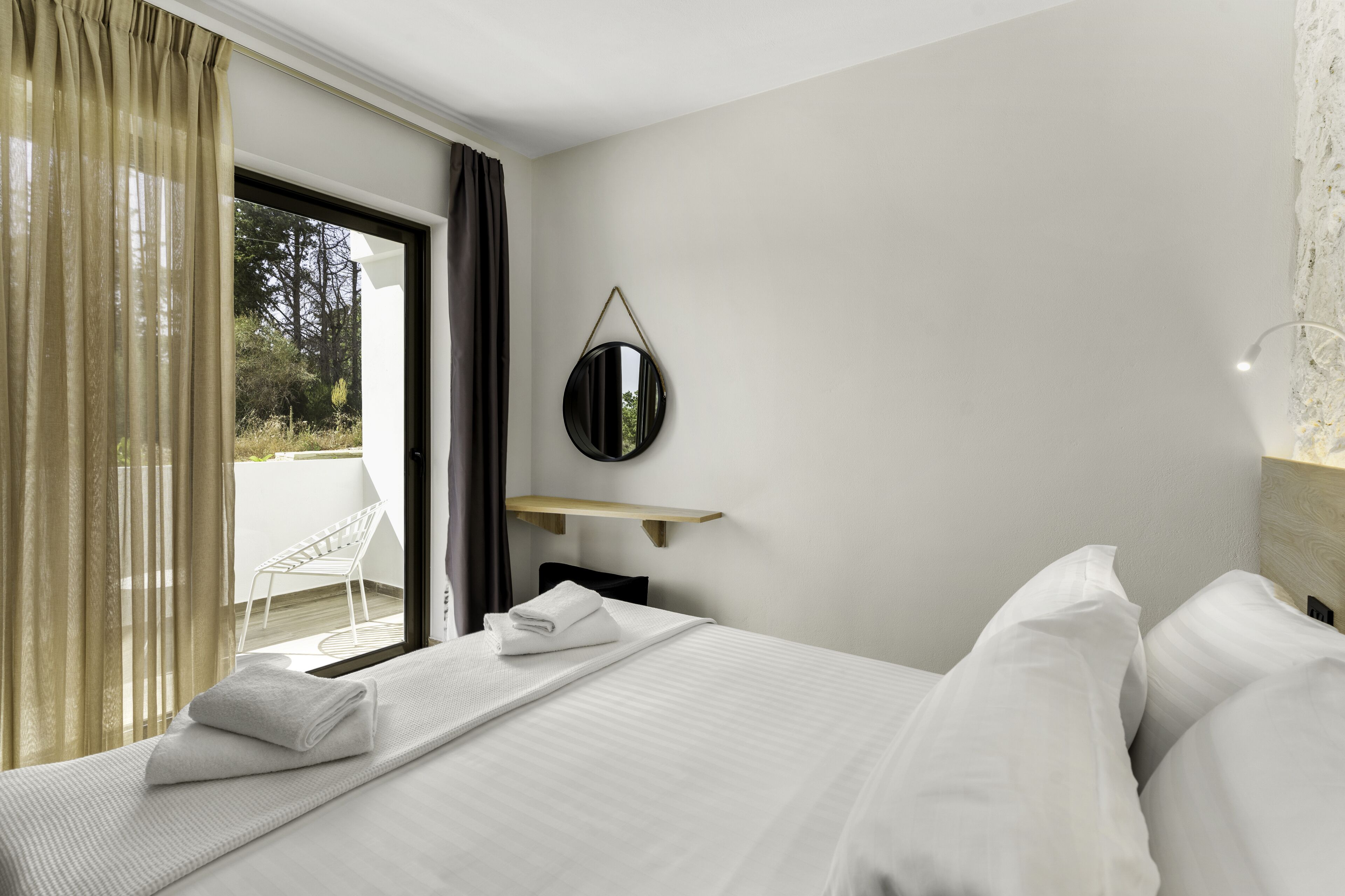 Habitación de hotel con una cama, un espejo redondo, un estante de madera pequeño, una silla blanca y vistas al exterior a través de una puerta de vidrio.