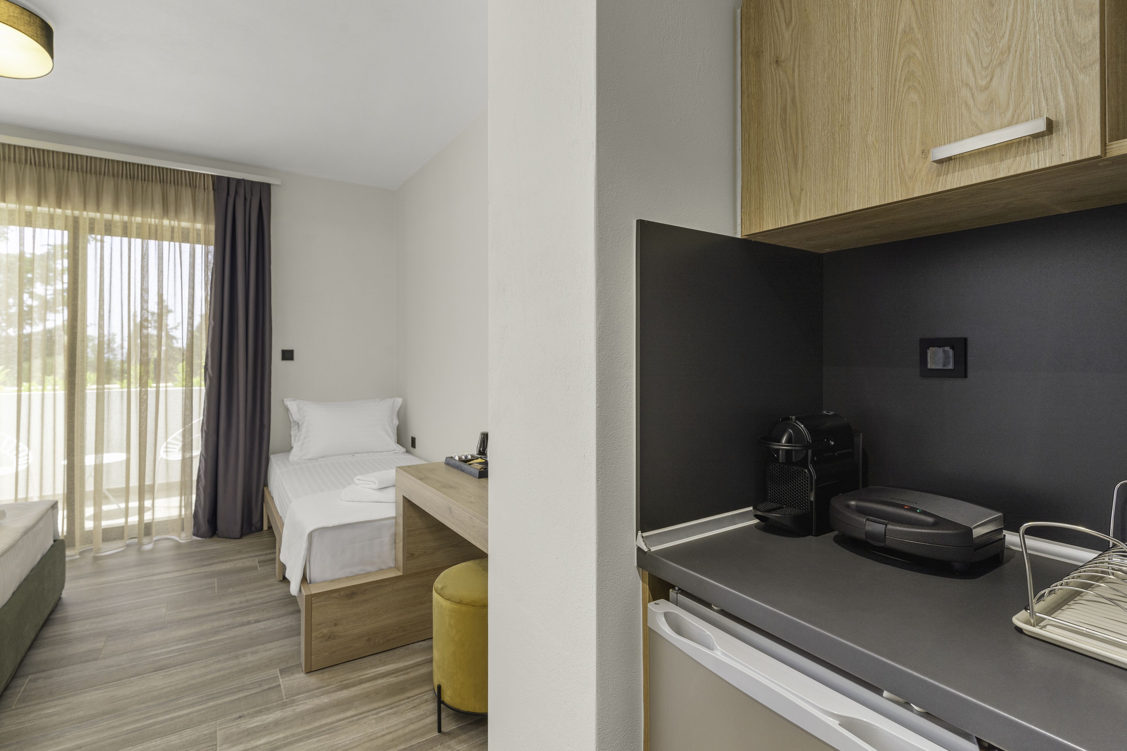 A modern hotel room with a bed, a small desk, a coffee maker, and a compact kitchen area.