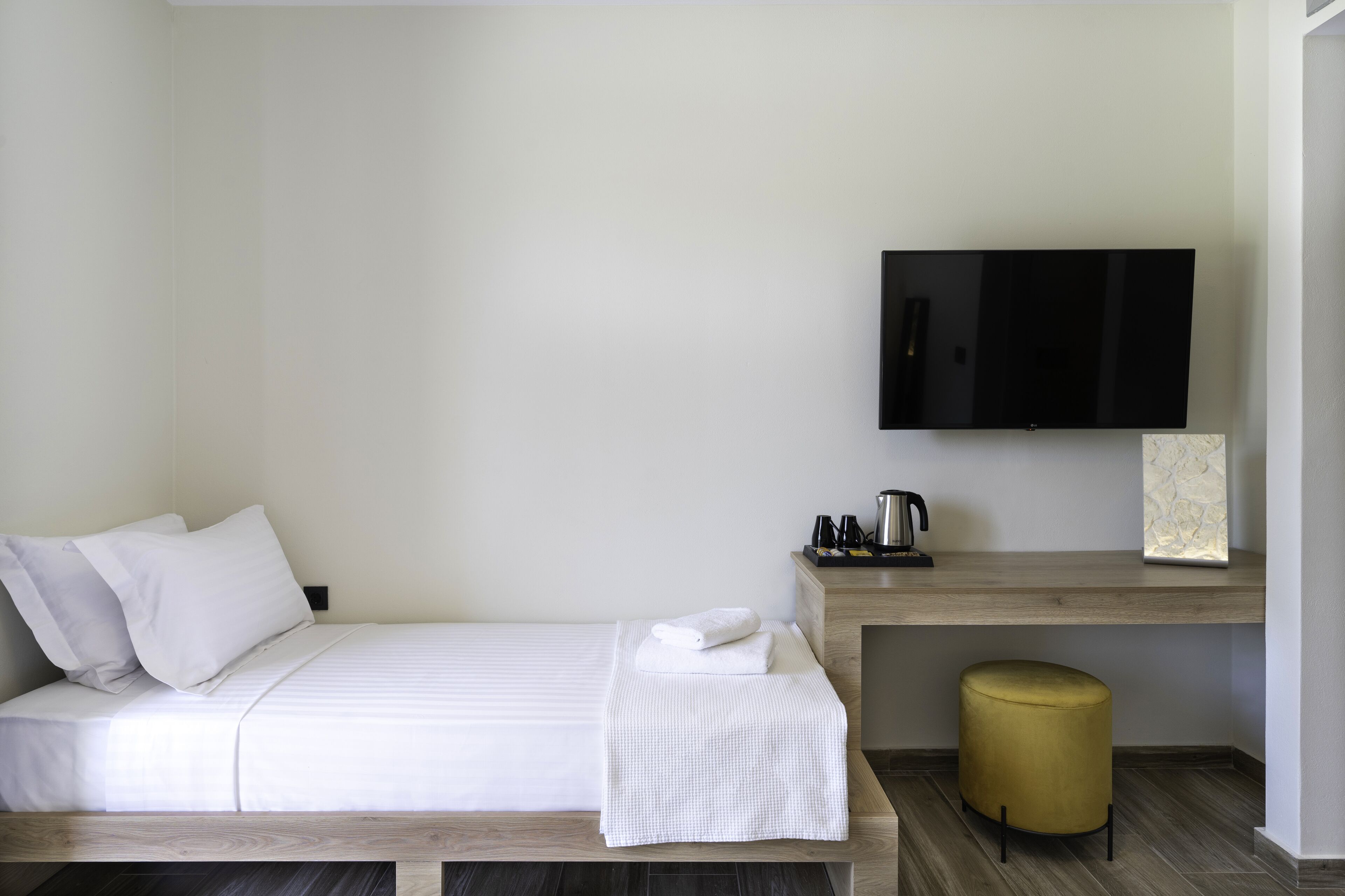 A neatly arranged hotel room with a bed, a flat-screen TV mounted on the wall, a small wooden desk with a coffee maker, and a round ottoman.