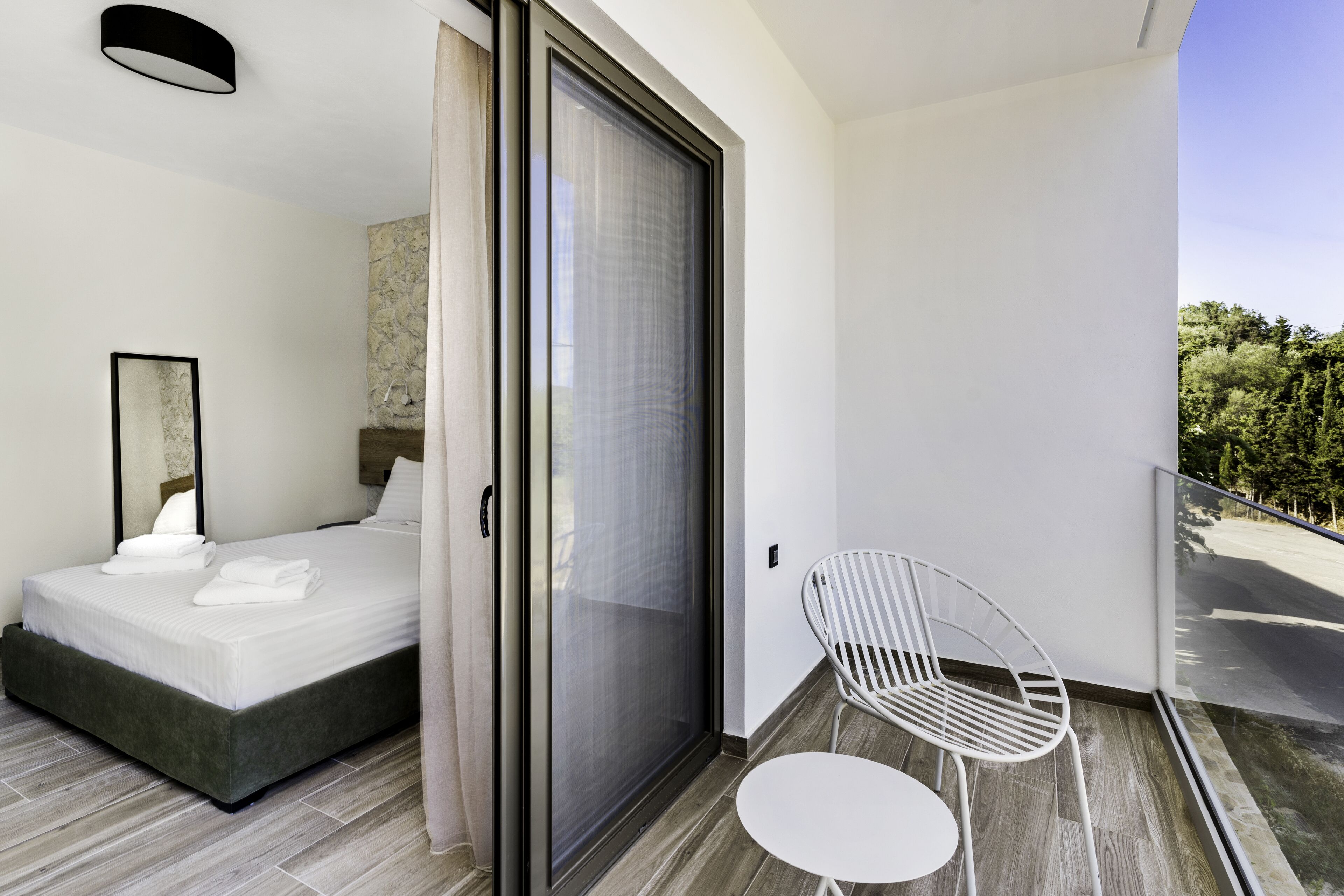 A modern hotel room with a bed, a chair, a small table, and a sliding glass door leading to a balcony.