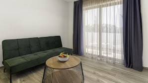 A room with a green sofa, a round wooden table with a bowl of fruit, and sheer curtains over a large window. - Sigma Luxury Rooms (Igoumenitsa)