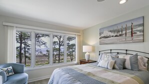 A bedroom with a large bed, a view of the sea, a painting of the sea, a chair, and a nightstand with a lamp. - Newly updated villa with pool access & stunning views of the Calibogue Sound! (Hilton Head Island)