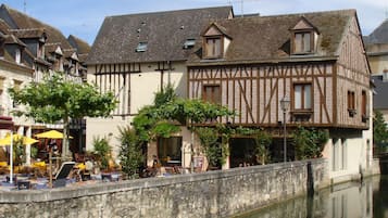 A traditional half-timbered building with a stone wall in front, a canal, and outdoor seating with umbrellas.