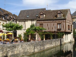 A traditional half-timbered building with a stone wall in front, a canal, and outdoor seating with umbrellas. - Moulin en Bord de Rivière, Logement Complet (Chevillon-sur-Huillard)