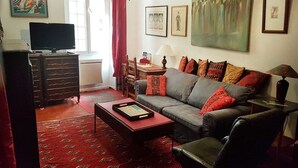 A living room with a grey sofa, red patterned cushions, a red carpet, a television on a wooden cabinet, and artwork on the walls. - Old Town Nice Family APT  Spacious  2BR;  A/C  3 min walk Beach cafes shops tram (Nice)