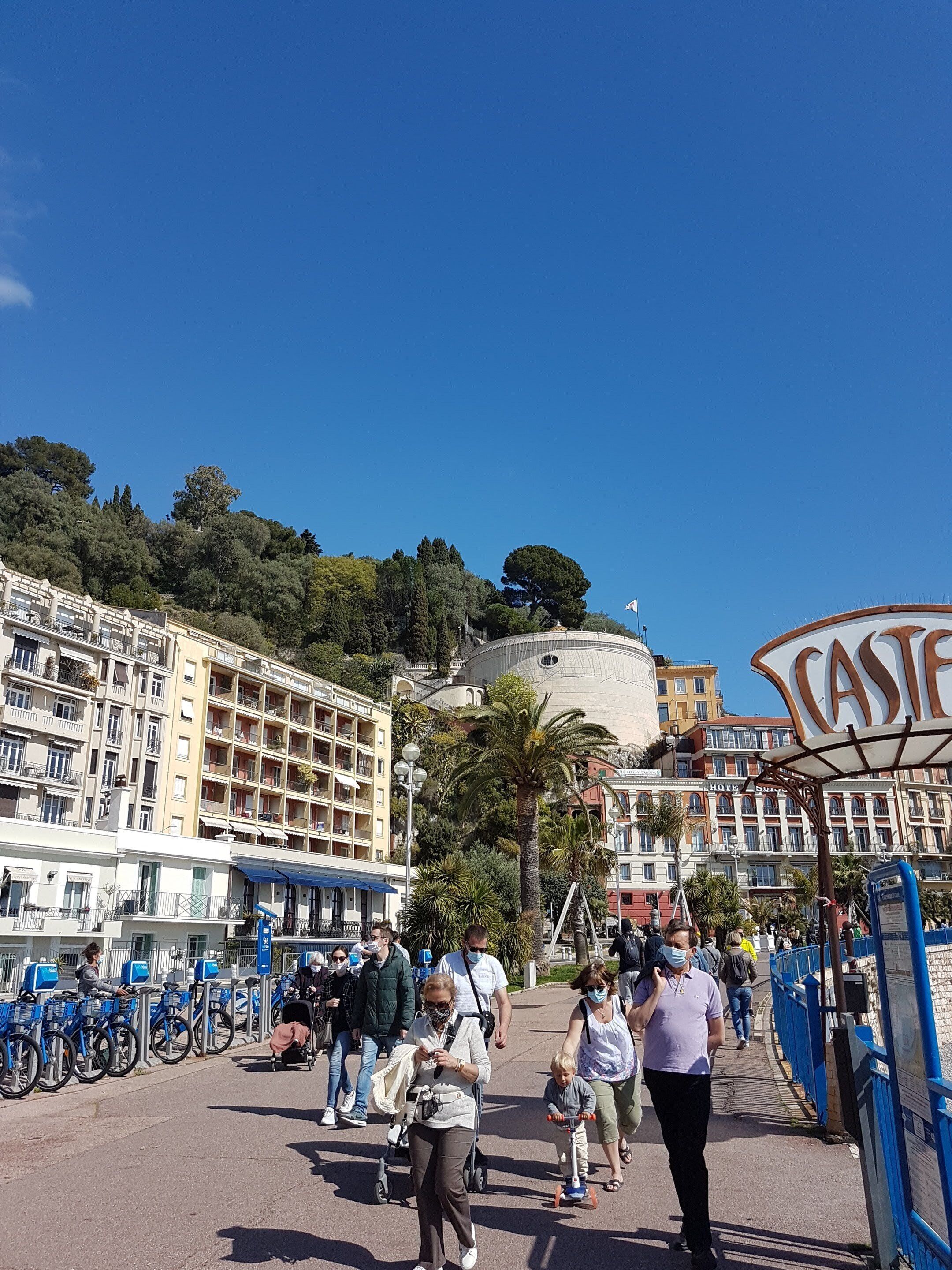 A busy promenade with people walking, a sign for 'CASTELLI', and a building with balconies and a dome.