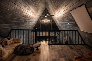 A cozy attic room with a wooden floor, a black railing, a chandelier, and a staircase.