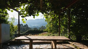 A stone patio with a wooden table and benches, surrounded by greenery and a view of distant mountains. - Vieiro Verde 2 Rustic (Vieiro)