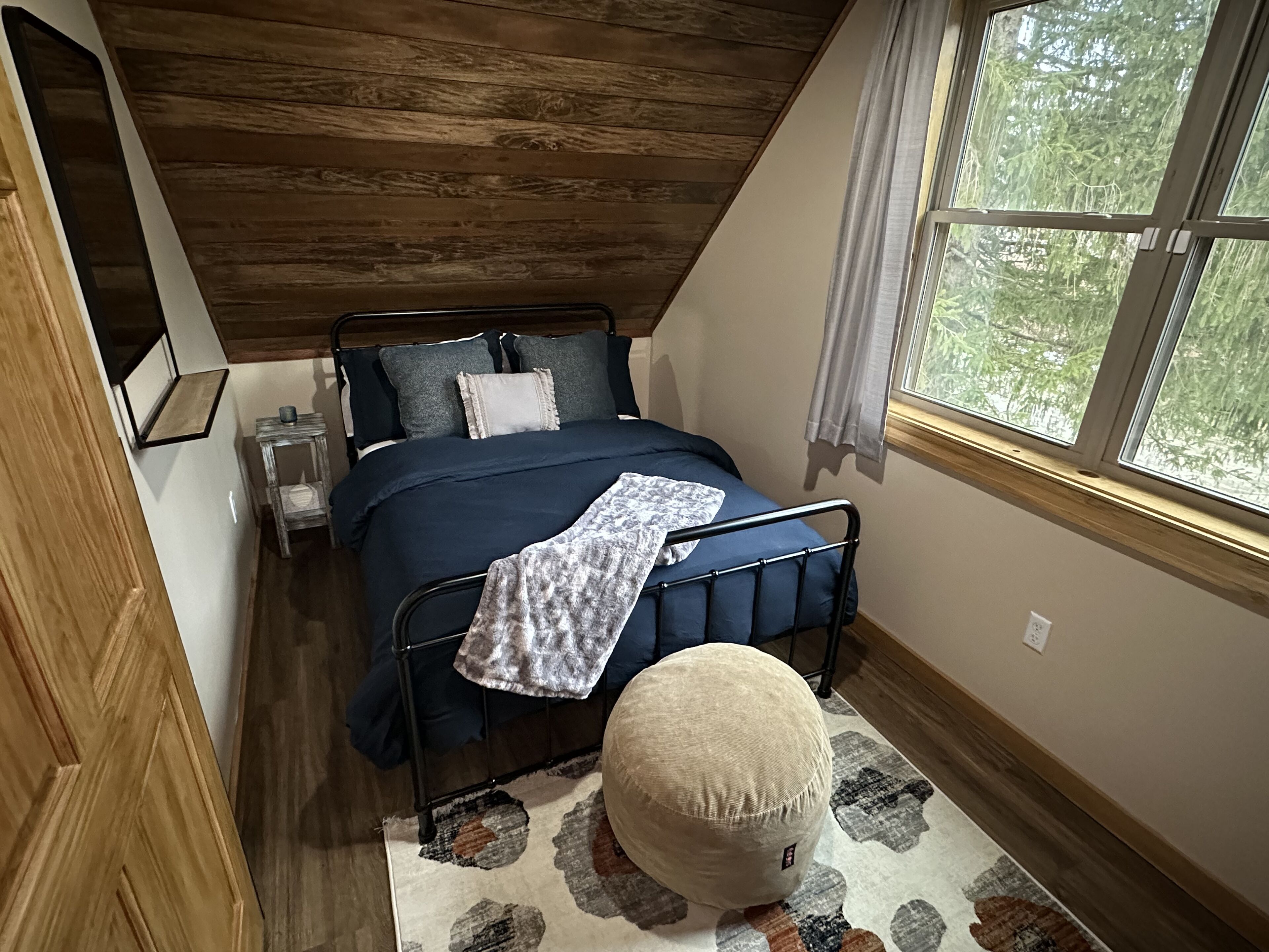 Ein Schlafzimmer mit schrägverdachter Decke, einem Bett mit blauer Bettdecke, einem Nachttisch mit Lampe und einem Fenster mit Blick auf Bäume.