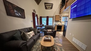 A modern living room with a sofa, armchair, and coffee table, a kitchen in the background, and a sign reading "Monongahela National Forest". - Parsons Riverside Retreat: Steps away from a rolling mountain stream and trails (Parsons)
