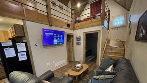A cozy living room with a black leather sofa, a small coffee table, and a TV mounted on the wall displaying a guest mode interface. - Parsons Riverside Retreat: Steps away from a rolling mountain stream and trails (Parsons)