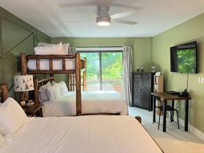 A bedroom with a bunk bed, a desk, a chair, a television, and a large window with a view of greenery.
