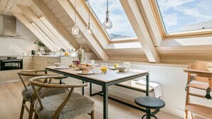 Um quarto moderno em estilo loft, com teto inclinado, mesa de jantar com cadeiras, cozinha e vista para as montanhas através de grandes janelas.
