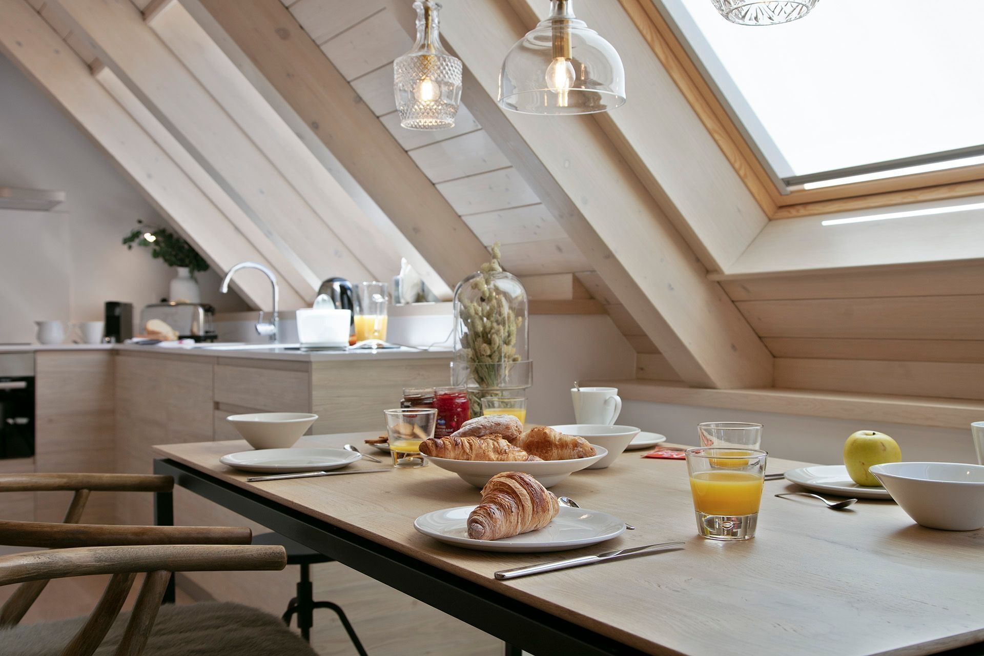 Une table de petit-déjeuner avec des croissants, du jus d’orange et un saladier de fruits, le tout disposé sur une table en bois dans une cuisine dotée d’une verrière.