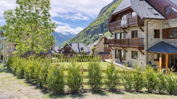 Um hotel em estilo chalé, com fachada de pedra, varandas de madeira e montanhas ao fundo.