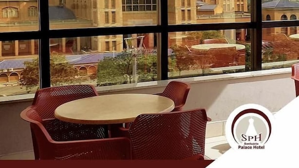 A room with large windows offering a view of a historic building with domes and arches. There is a round table with chairs and a hanging light fixture. The logo of the Santuário Palace Hotel is visible.
