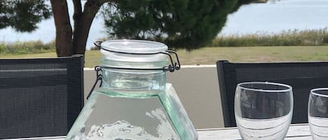 A table with a glass jar, a wooden mug, and a glass cup, set outdoors with a view of a tree and water.