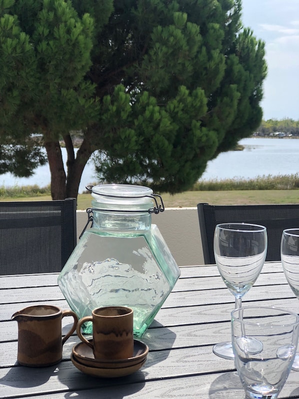 A table with a glass jar, a wooden mug, and a glass cup, set outdoors with a view of a tree and water.
