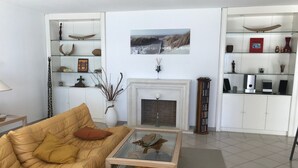 Una sala de estar con un sofá beige, una mesa de centro de vidrio, una chimenea y estantes con objetos decorativos y libros.