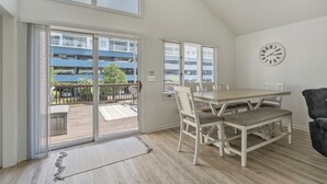 Un área de comedor con una mesa y sillas de madera, un reloj en la pared y vistas a un edificio azul a través de amplios ventanales.