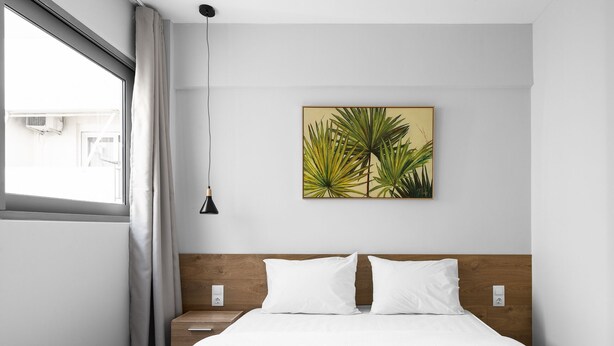 Ein Hotelzimmer mit Bett, weißen Leinen, zwei Kissen, einem Kopfteil, einem Wandbild, einem Nachttisch, einer Lampe und einem fenster mit Vorhängen.