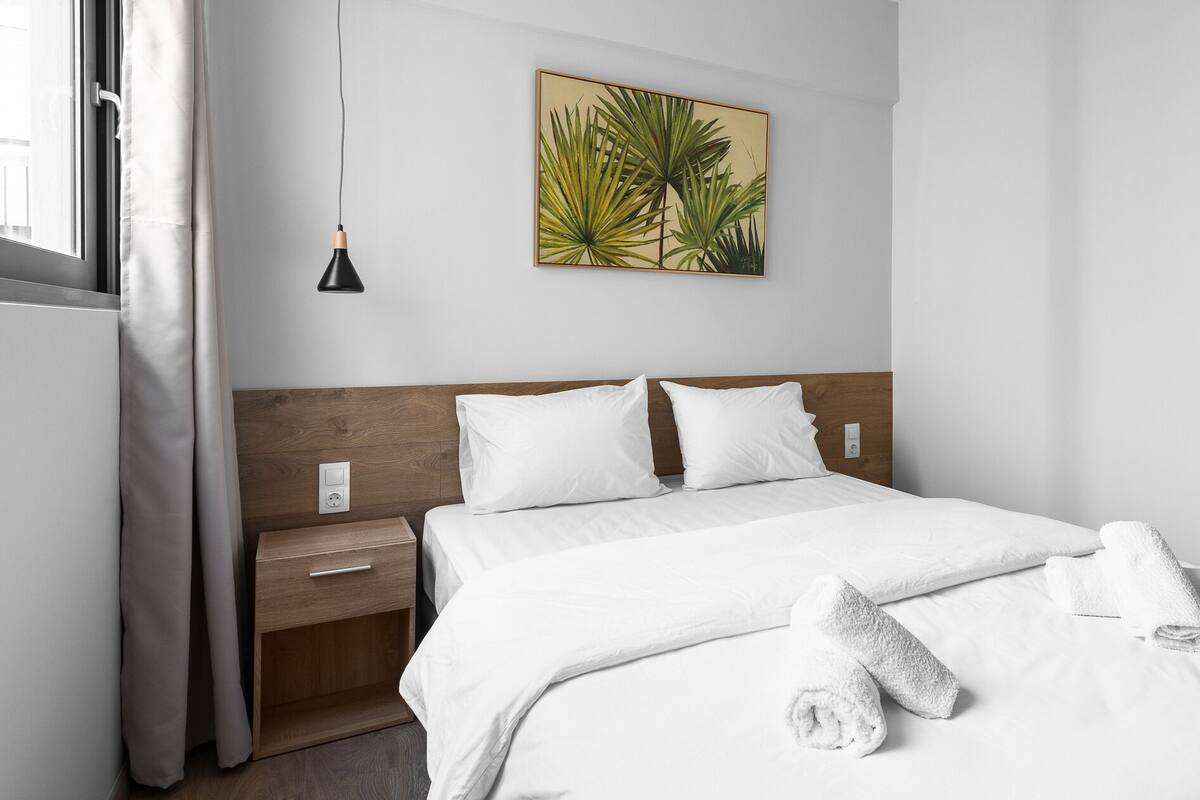 A hotel room with a wooden headboard, white bedding, a bedside table with a drawer, a wall-mounted painting of a plant, and a hanging pendant light.