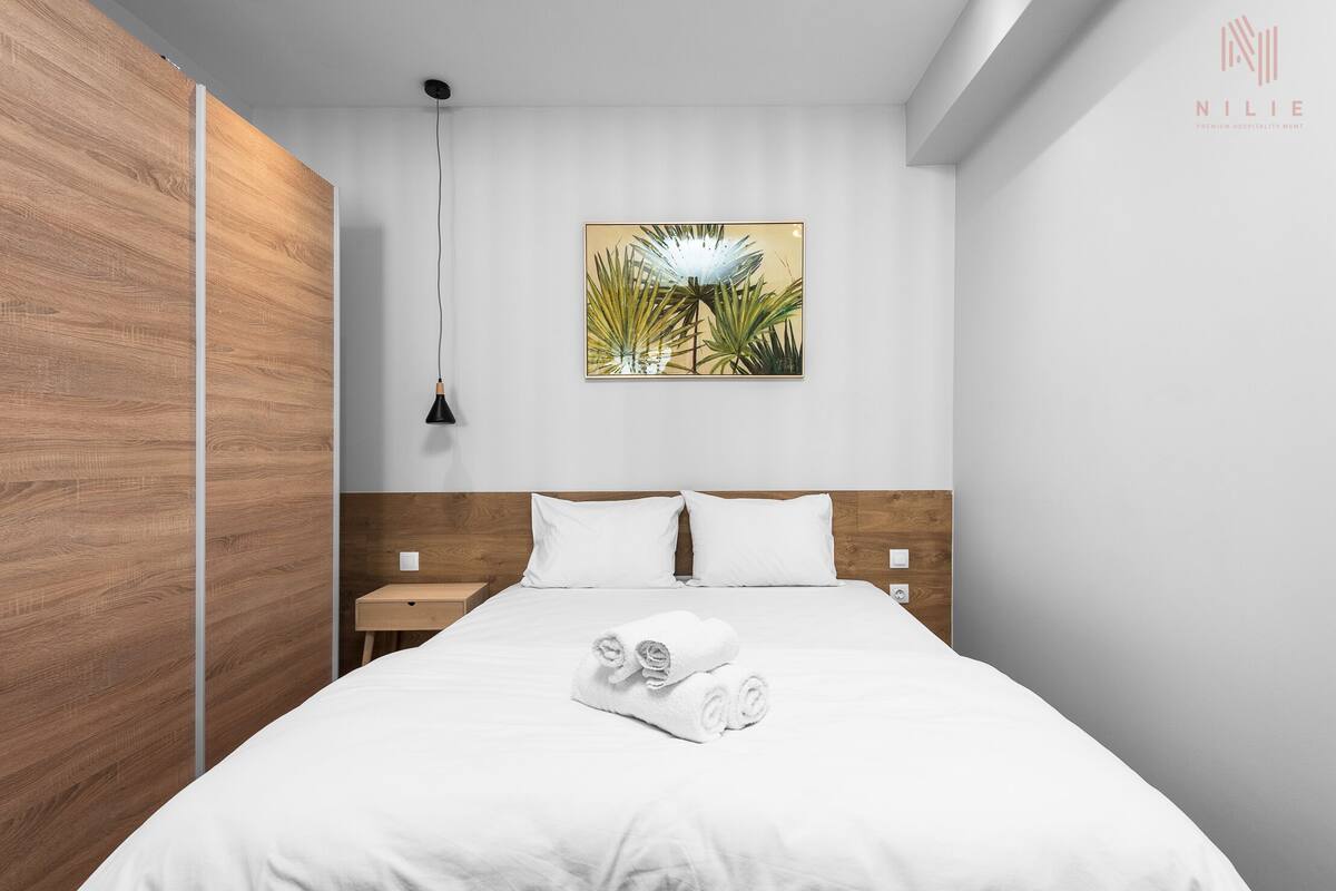 A modern hotel room with a wooden accent wall, a bed with white linens, a nightstand, a wall-mounted lamp, and a tropical-themed painting.